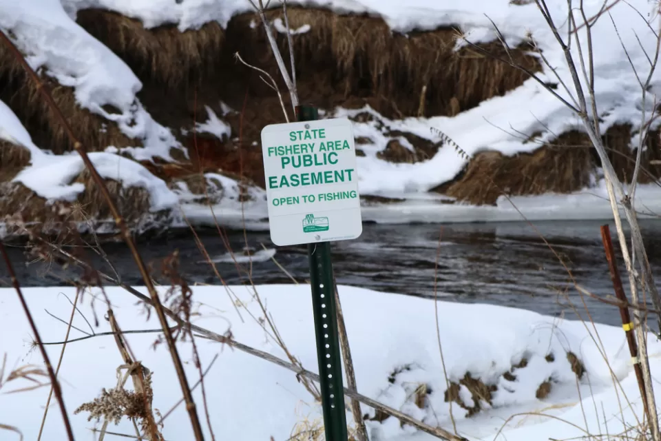 A sign reads “State Fishery Area Public Easement Open to Fishing.” In the background is a stream surrounded by snow on the ground in a natural setting.